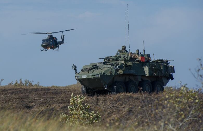 A light armoured vehicle is in a field while a military helicopter flies above it.