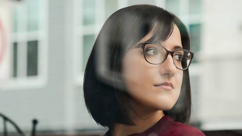 A woman wearing glasses holds a mug inside a coffee shop and looks out the window to her right.