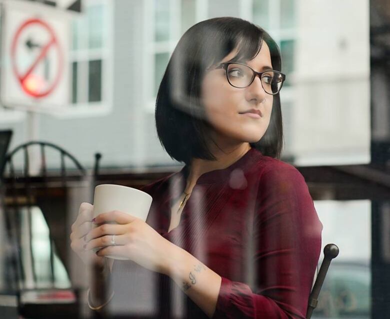 A woman wearing glasses holds a mug inside a coffee shop and looks out the window to her right.