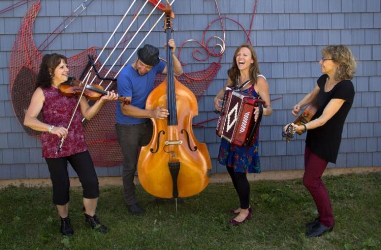 Four musicians play fiddle, stand-up bass, accordion, and guitar, outside in front of a wooden building. 