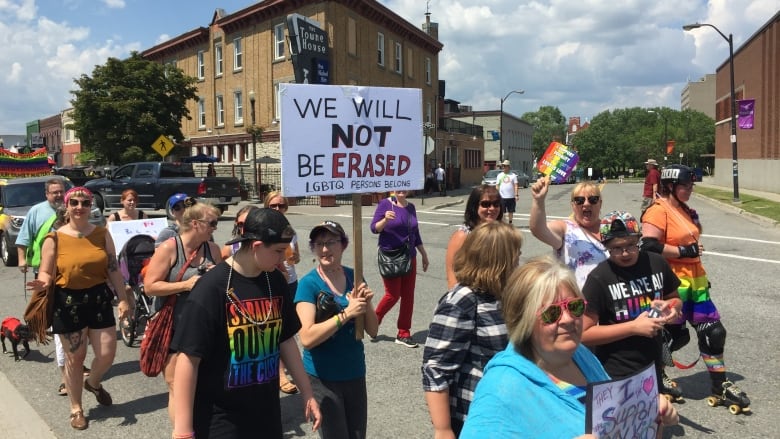 A group of people marching along a city street. One person is carrying a sign that says we will not be erased.