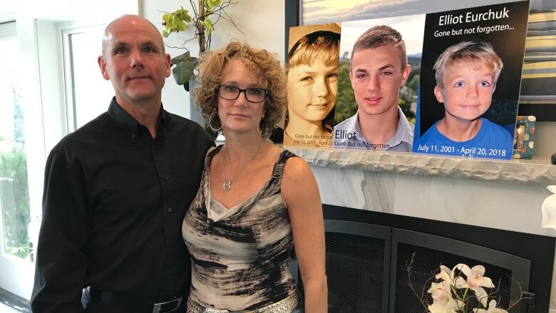 A man and a woman stand in front of a fireplace with pictures of their son displayed above the mantle.