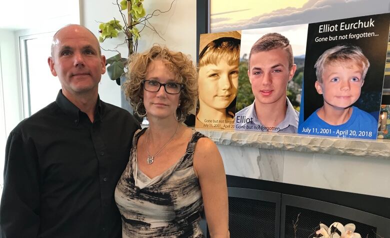 A man and a woman stand in front of a fireplace with pictures of their son displayed above the mantle.