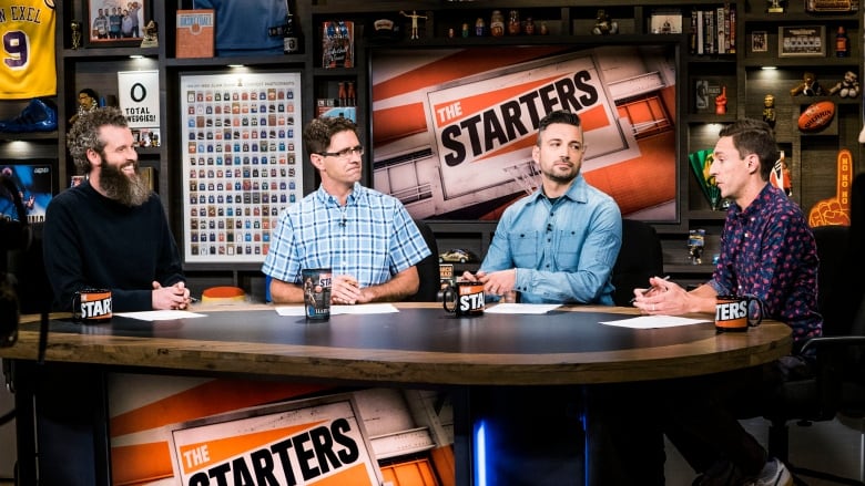 Four people sit around a table on a TV set. 