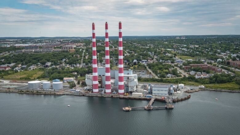 Three red and white striped towers of a power generating plant stand at the edge of a harbour. Trees and houses can be seen in the distance.