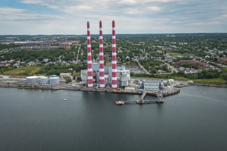 Three red and white striped towers of a power generating plant stand at the edge of a harbour. Trees and houses can be seen in the distance.