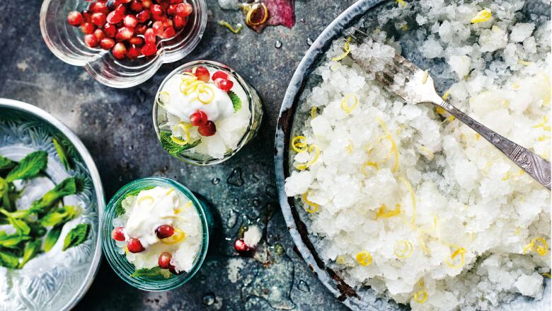 overhead shot of a grey plate of shaved ice with lemon rind in it. 2 small glasses of the dessert topped with cream and pomegranate arils sit next to it, all on a dark grey surface. 