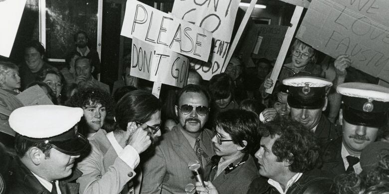 In a scene from a film, a smiling man in sunglasses is surrounded by supporters and police, with a placard saying 