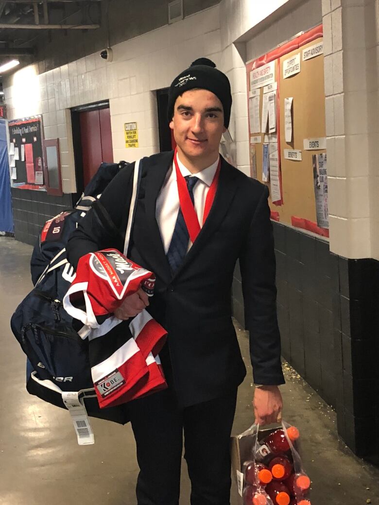 A young man in a black suit holds his hockey jersey and bag in the middle of a hallway.