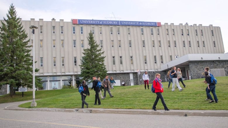 Some students walk on the grass in front of a grey institutional building with a sign reading 'University of Sudbury' 