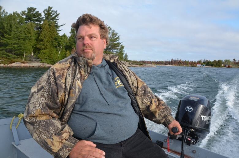 A man with a beard, wearing a camouflage jacket, drives a boat down a river 