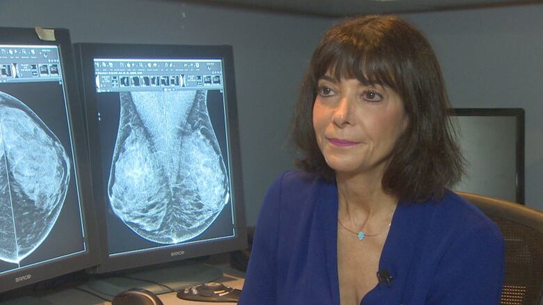 A woman with shoulder-length brown hair, wearing a blue dress, sits in front of mammogram images on a screen