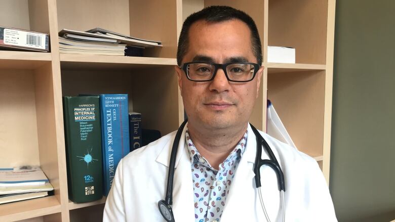 Dr. Kym Jim wears a white lab coat and stethoscope around his neck as he sits in front of a book shelf in his office.