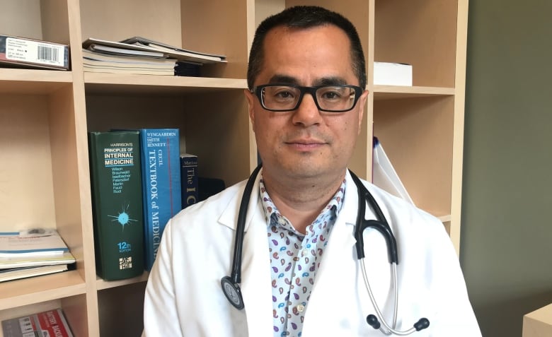 Dr. Kym Jim wears a white lab coat and stethoscope around his neck as he sits in front of a book shelf in his office.