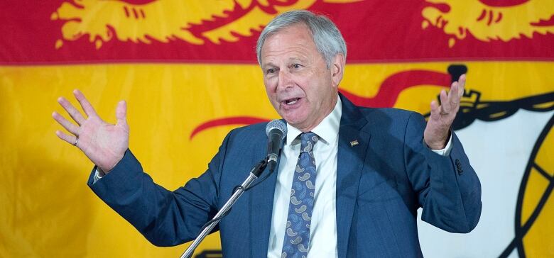 Premier Blaine Higgs speaks in front of a flag of New Brunswick.