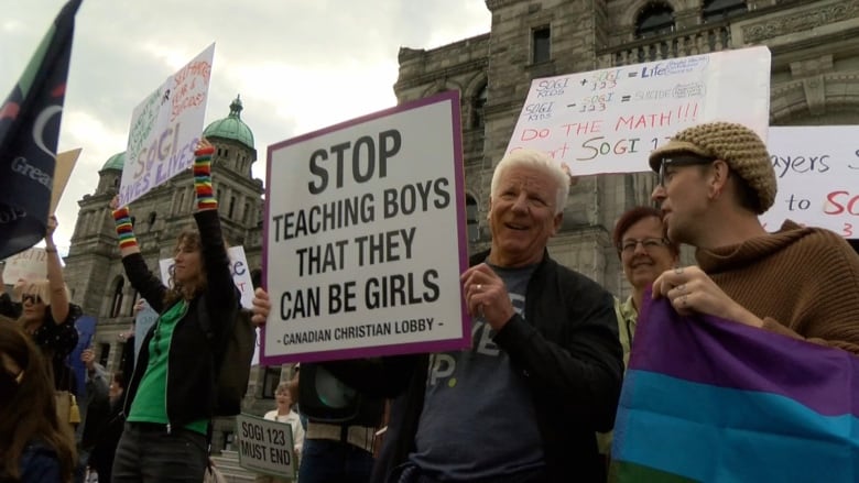 People hold signs both for and against SOGI, one reading 'Stop teaching boys that they can be girls'