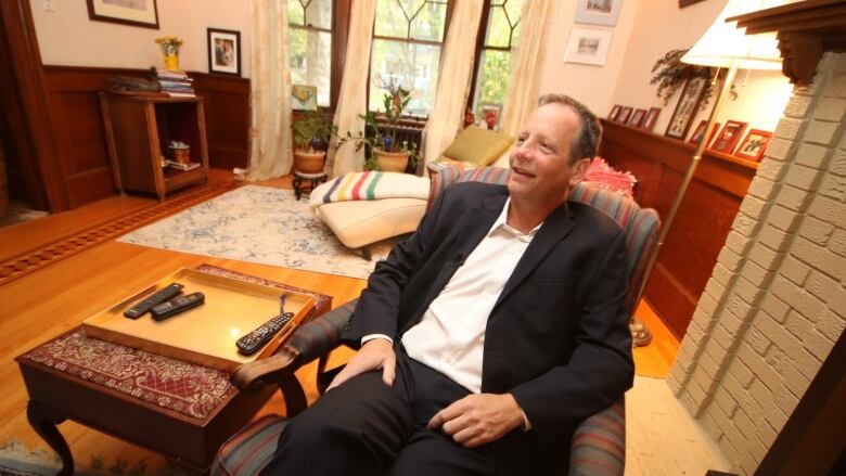 A man is sitting in a chair in a living room, with couches and tables in the background.