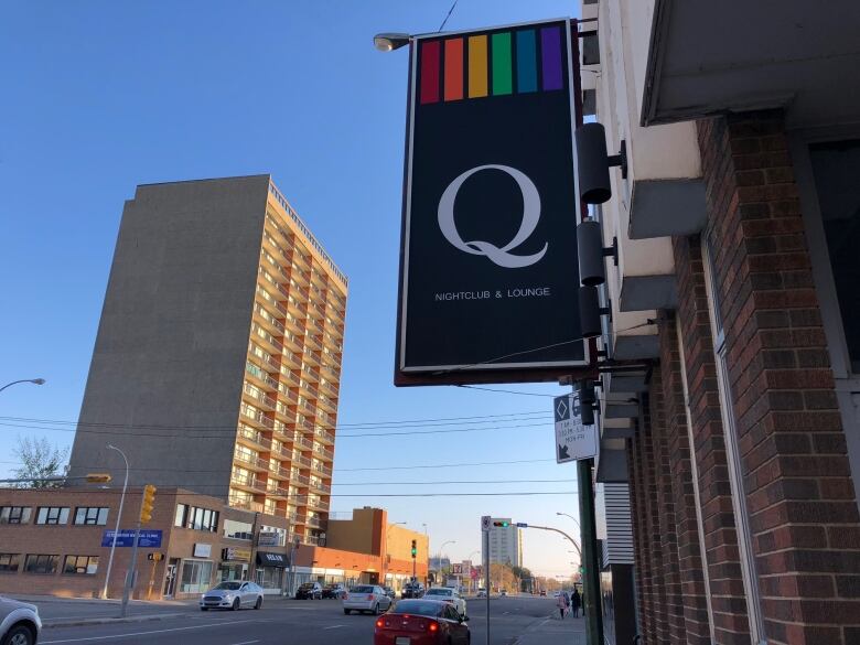 A black sign topped with vertical stripes in rainbow colours and featuring a large white letter Q is attached to the side of a brick building on an urban street.