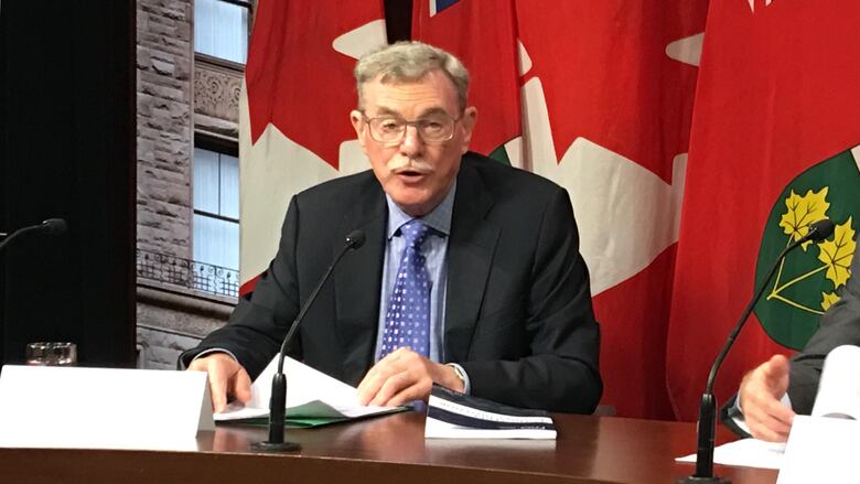 Photo of David Wake seated at a desk in the media studio at Queen's Park, with flags in the background. 