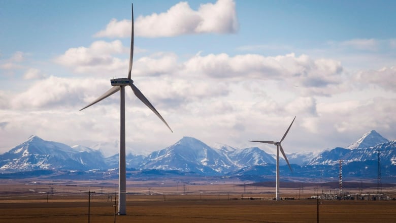 Two large wind turbines are pictured on rural pasture land with the Rocky Mountains in the distance against a partly cloudy sky.