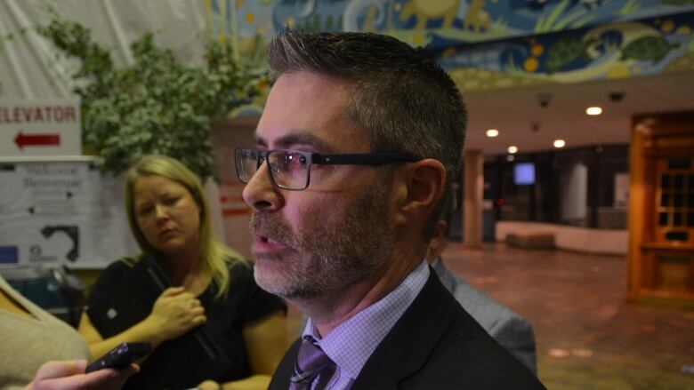 Close up photo of a man speaking into several microphones, with a reporter listening in the background. 