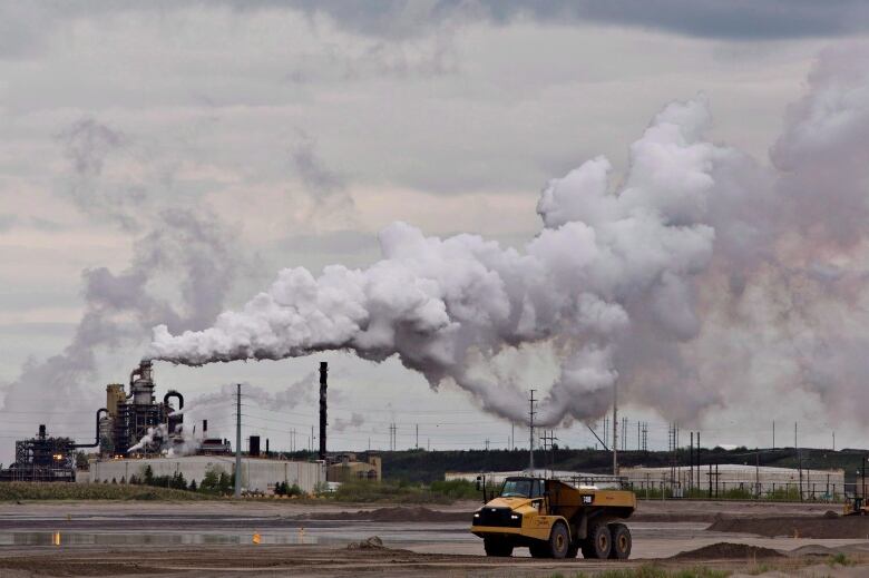 A dump trump stops in front of a large oil sands extraction facility. A large plume of smoke billows rightwarsd from a column.