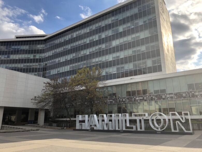 A large glass building with a sign reading HAMILTON in front is seen on a sunny day