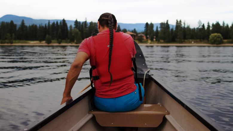 A man paddles a canoe toward a forest in the distance.