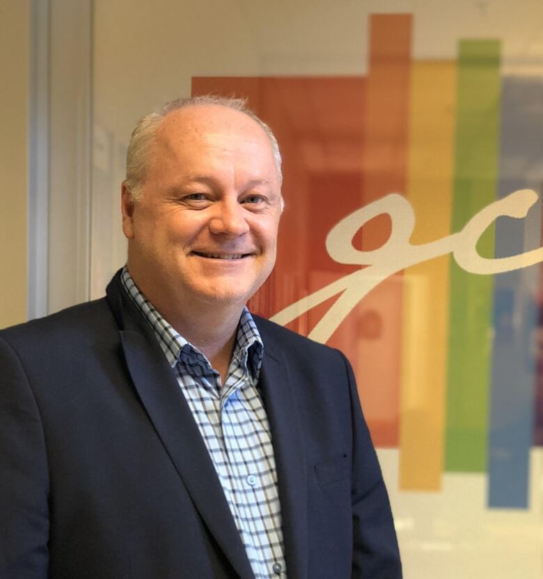 Jason Maclennan smiles widely wearing a solid navy suit jacket with a checked, collared shirt underneath. There are rainbow colours on the wall behind him.