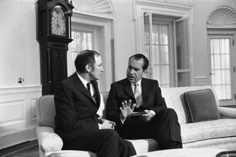 Two men talk while sitting on a couch in black-and-white photo