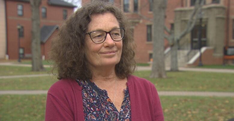 A woman with long brown hair and glasses stands on a university campus.