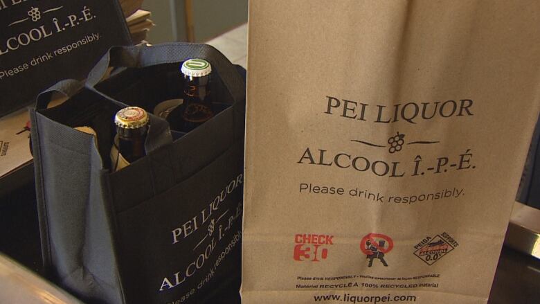 Paper bags with P.E.I. Liquor Control Commission branding sits on the conveyor belt of a check out counter. A cloth bag sits beside it with glass bottles inside. 