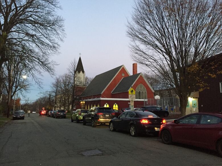 A red church building is seen around dusk. There is a line of cars at the front.