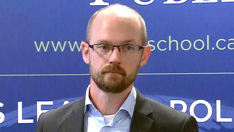 Trevor Tombe is seen wearing a suit jacket and a blue-collared shirt. He's standing in front of a blue backdrop.