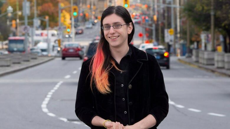 A woman in black with glasses and pink highlights in her hair smiles with an urban streetscape in the background.