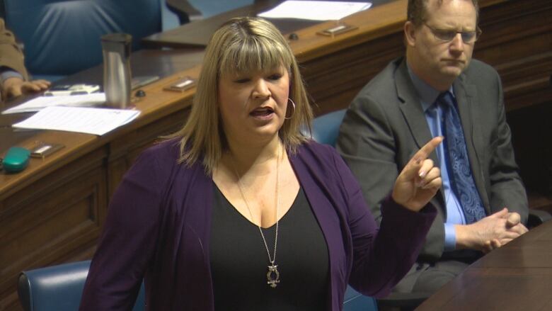 A woman with long brown hair and wearing a purple cardigan over a black shirt, gestures with her hands as she speaks in a room with other people sitting nearby.