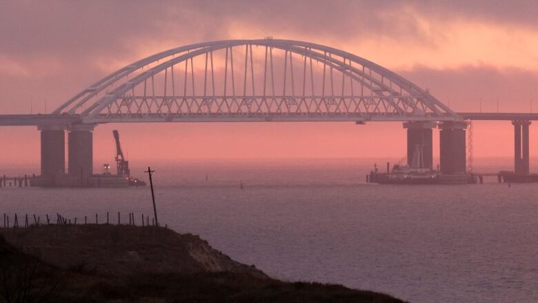 A long bridge is shown spanning waters against a pink sky at dusk.