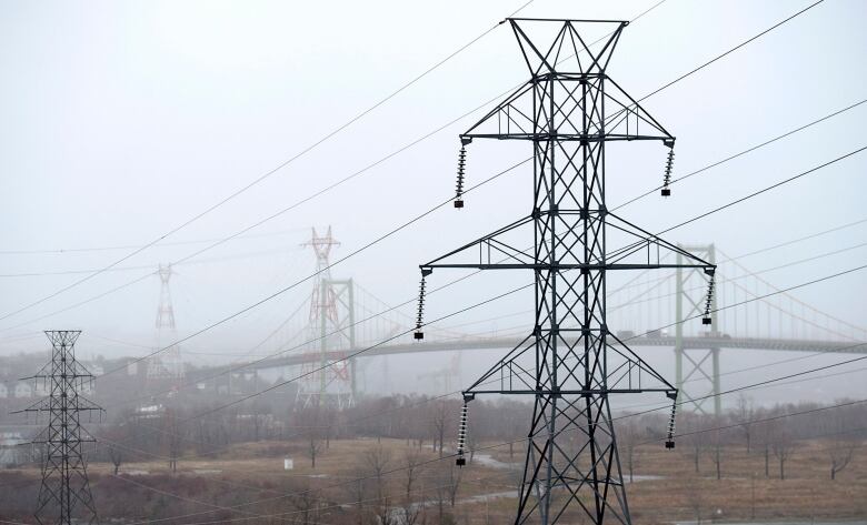 Power line towers stand in a field, with a bridge in the background, on a foggy day.