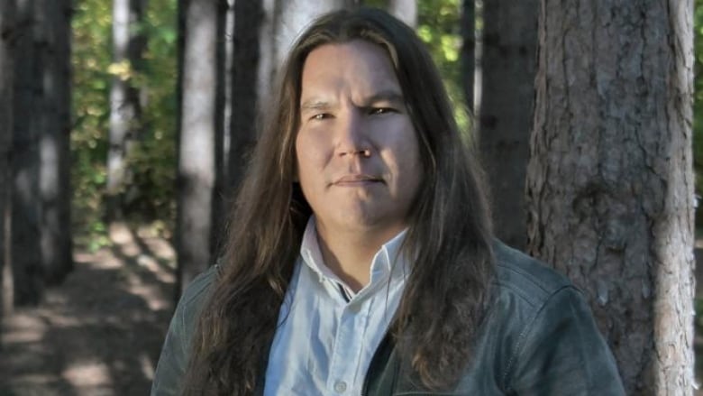 Close up portrait of a man with long dark hair wearing a light blue shirt with a dark grey jacket over it and standing in front of a tree