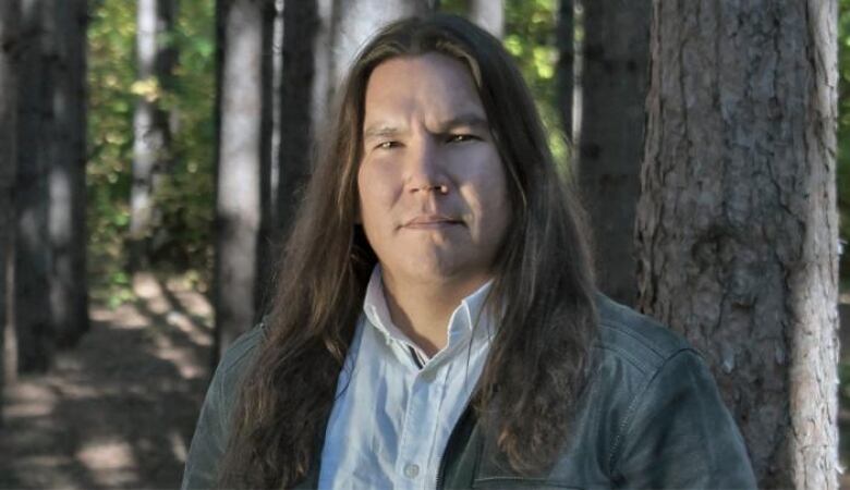 Close up portrait of a man with long dark hair wearing a light blue shirt with a dark grey jacket over it and standing in front of a tree