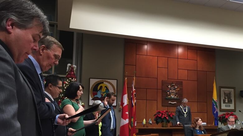 Several people in dresses and suits stand and read an oath, some with their hands up or over their hearts, in a council chambers. 