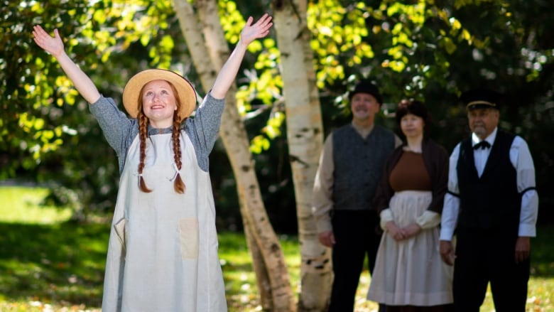 A young redhaired girl in early 19th-century clothing, wearing a straw hat and apron, raises her arms into the air. 