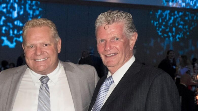 Two middle-aged white men smile with an arm around each other's shoulder, posing for the camera. One is the premier of Ontario. 