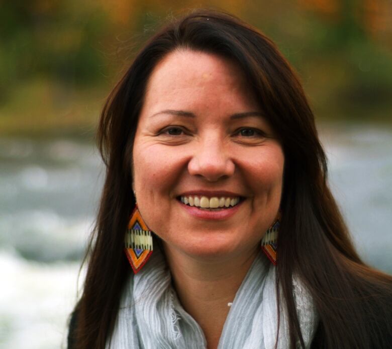 A woman stares into the camera with a smile wearing a scarf with water and trees in the background.