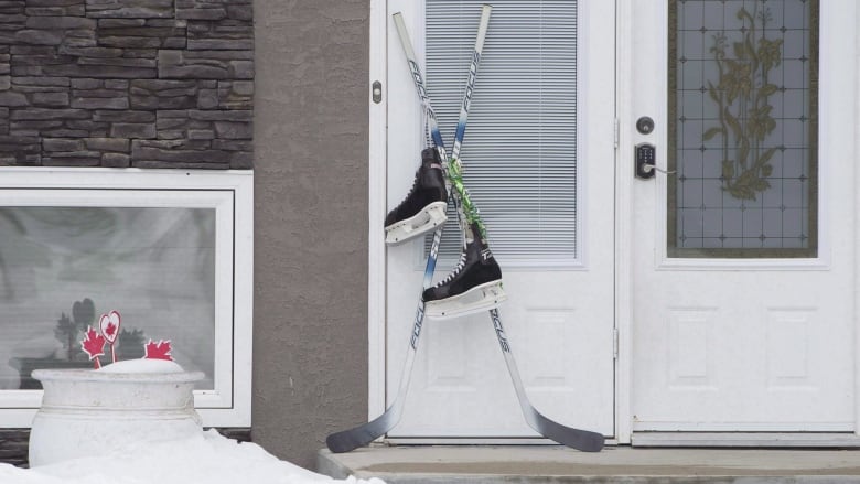 A pair of hockey sticks are set on a front door in 2018. They are crossed and a pair of skates hangs from them. 