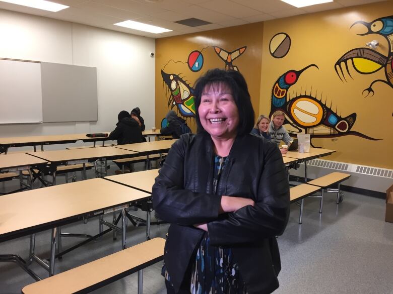 Margaret stands in what appears to be an almost empty school lunch room with bright woodland art painted in a mural across one wall.  A few kids are at tables in the back near the wall.  Margaret has a huge small on her face. 