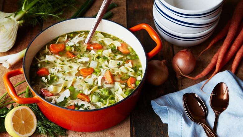 A red Dutch oven filled with chicken noodle soup, sitting on a wooden table with carrots, onions, soup bowls and spoons sitting next to it. 