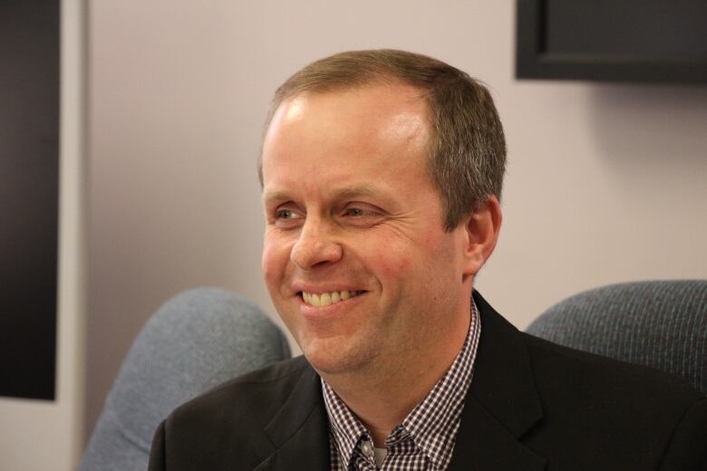 A man with a checked shirt and dark suit jacket smiles.