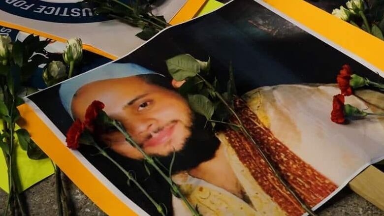 A picture of a man smiling at the camera, pasted on paper on top of a stick. Flowers are seen on top of the photo.Soleiman Faqiri was born on New Year's Day in Kabul, Afghanistan, in 1986 and came to Canada in 1993. According to his family, he was a straight-A student, captain of his high school rugby team and had a close and loving relationship with his four siblings and parents. 