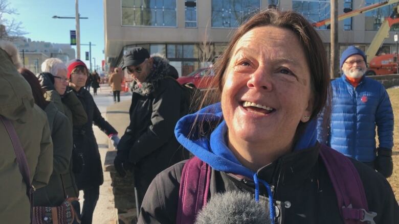 A woman with a ponytail and a blue sweater under a black jacket smiles at the camera while standing on a sidewalk